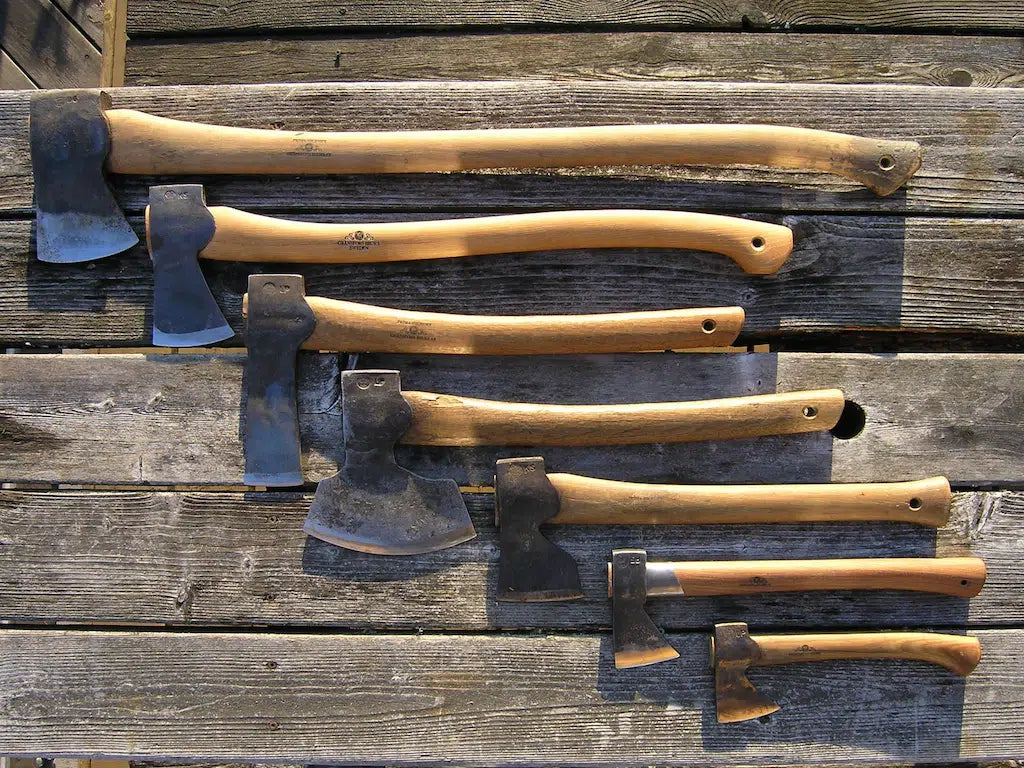 Six hand axes with light brown wooden handles and dark steel heads arranged horizontally on a rustic weathered wood plank surface.